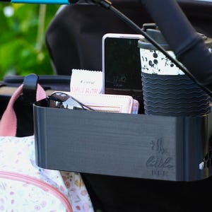 Può includere: Un organizer per passeggino nero con portabicchieri, telefono, portafoglio e occhiali da sole. L'organizer ha il testo "The Little Bee" ed è attaccato a un manico del passeggino. Un bicchiere da viaggio con un design in bianco e nero è nel portabicchieri.