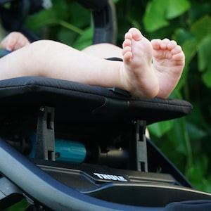 Può includere: Primo piano dei piedi di un bambino appoggiati sul sedile nero di un passeggino. Il passeggino ha un telaio nero e il marchio "THULE" è visibile. Fogliame verde sullo sfondo.