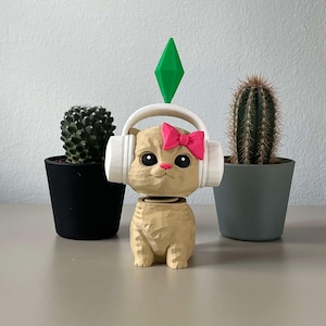 Pode incluir: Estatueta de gato bege com fones de ouvido brancos e laço rosa, com um diamante verde em cima. Dois cactos em vasos ladeiam o gato em uma superfície neutra.