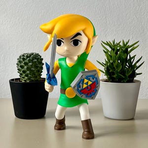 Op de afbeelding: Een beeldje van Link uit The Legend of Zelda, in een dynamische pose. Hij draagt een groene tuniek, geel haar en een schild met een rood wapen. Hij houdt een blauw zwaard vast, klaar voor actie. De figuur is ongeveer 13 cm hoog.