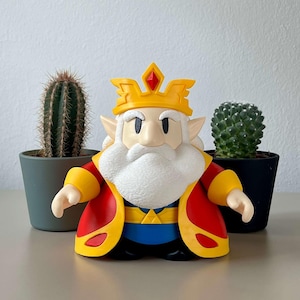Puede incluir: Una colorida figura de un rey con corona amarilla y túnica roja. La figura tiene barba blanca y está frente a dos cactus en macetas. La túnica del rey tiene ribetes amarillos y una camiseta interior azul.