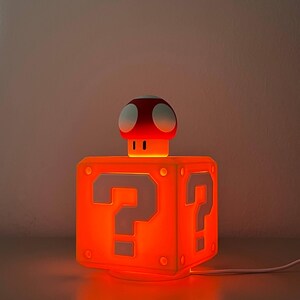 May include: A glowing orange cube-shaped lamp featuring a white question mark on each side. A small red and white mushroom-shaped figure sits on top of the cube. The lamp emits a warm orange light and includes a white power cord extending from the base.
