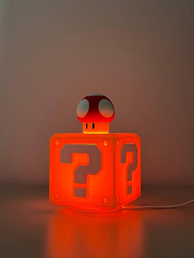 May include: A glowing orange cube-shaped lamp featuring a white question mark on each side. A small red and white mushroom-shaped figure sits on top of the cube. The lamp emits a warm orange light and includes a white power cord extending from the base.