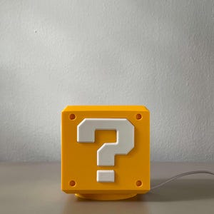 May include: A yellow cube-shaped lamp featuring a white question mark symbol on the front face. The design includes four small circular indentations at the corners of the front panel and a thin gray power cord extending from the right side of the base.
