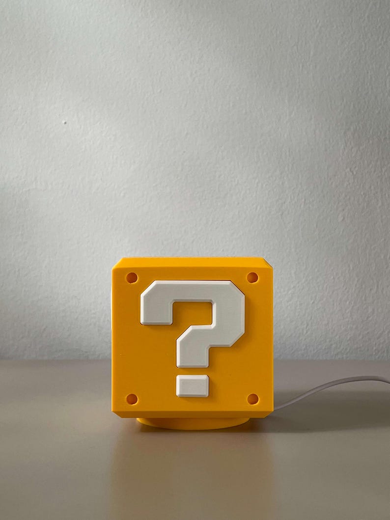 May include: A yellow cube-shaped lamp featuring a white question mark symbol on the front face. The design includes four small circular indentations at the corners of the front panel and a thin gray power cord extending from the right side of the base.