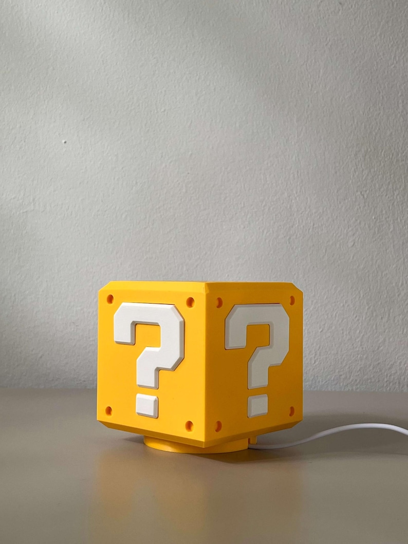 May include: A yellow cube-shaped lamp featuring a white question mark symbol on each visible side. The lamp has a structured design with small circular indentations at the corners and is connected to a white power cord extending from the base.