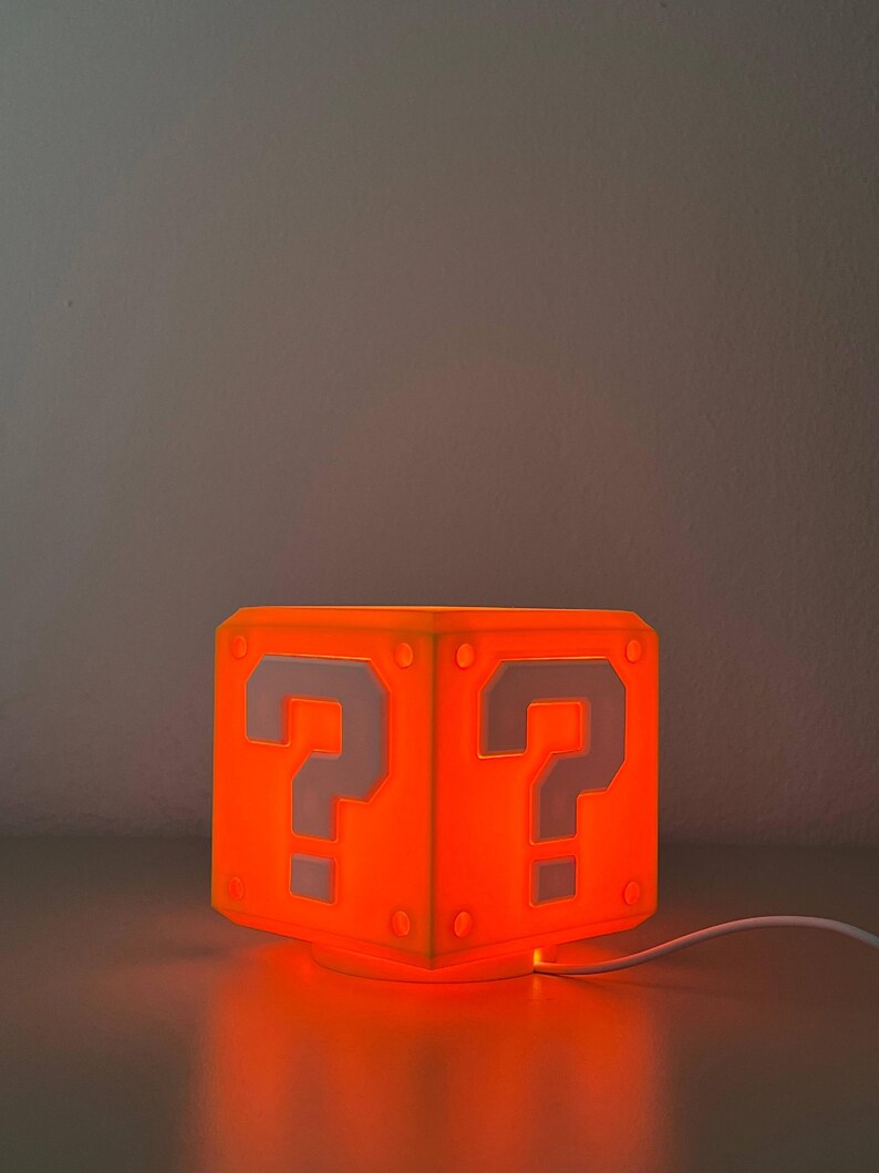 May include: A cube-shaped lamp emitting a bright orange glow. The exterior features a white question mark symbol on each visible side, framed by a raised border with circular corner accents. A white power cord extends from the base of the lamp.
