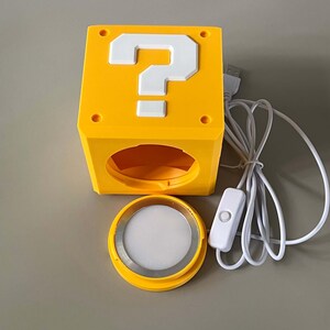 May include: A yellow cube-shaped lamp featuring a white question mark on the top face. The lamp has a circular opening on the front side, a separate circular light component with a white diffuser, and a white USB power cord with an inline switch.