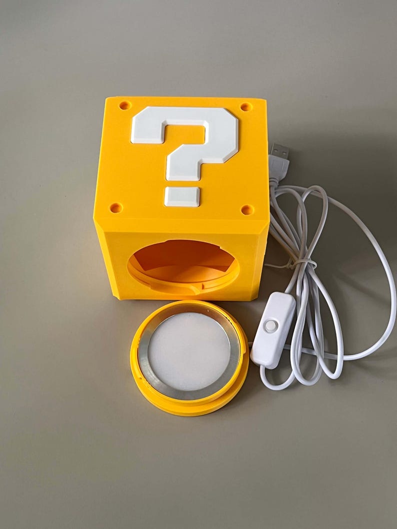 May include: A yellow cube-shaped lamp featuring a white question mark on the top face. The lamp has a circular opening on the front side, a separate circular light component with a white diffuser, and a white USB power cord with an inline switch.