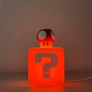 May include: A square orange lamp featuring a prominent question mark symbol on the front face. A small mushroom-shaped figure with a red cap and white spots sits atop the block. The entire structure emits a warm, bright orange glow.