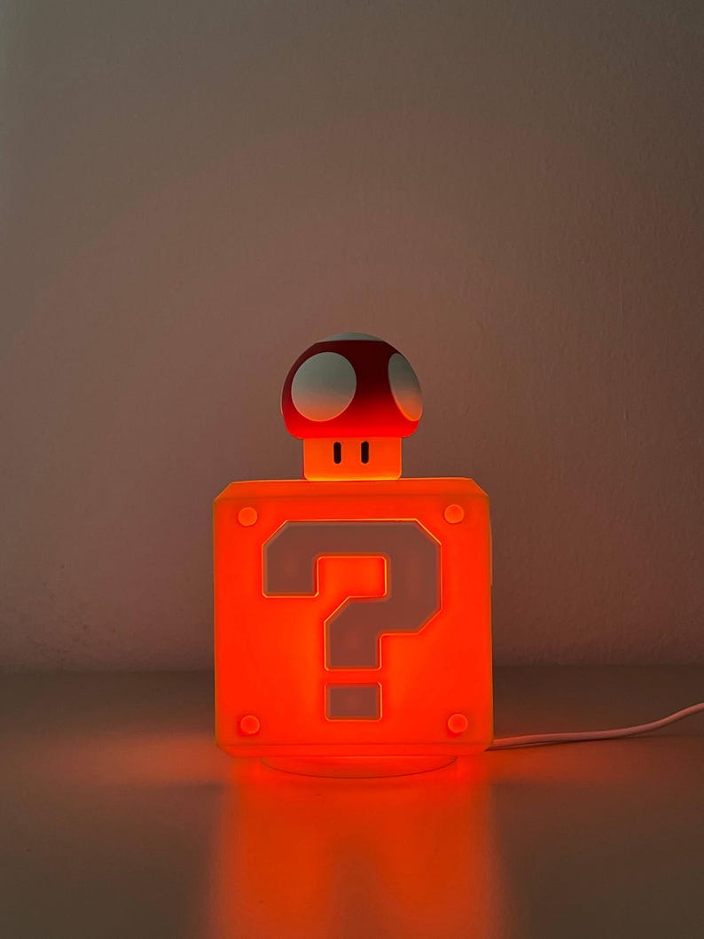 May include: A square orange lamp featuring a prominent question mark symbol on the front face. A small mushroom-shaped figure with a red cap and white spots sits atop the block. The entire structure emits a warm, bright orange glow.