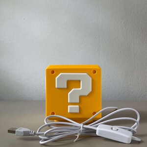May include: A yellow cube-shaped lamp featuring a white question mark symbol on the front face. The lamp includes a white power cord with a USB connector and an inline toggle switch for operation, designed with a smooth, matte finish surface.