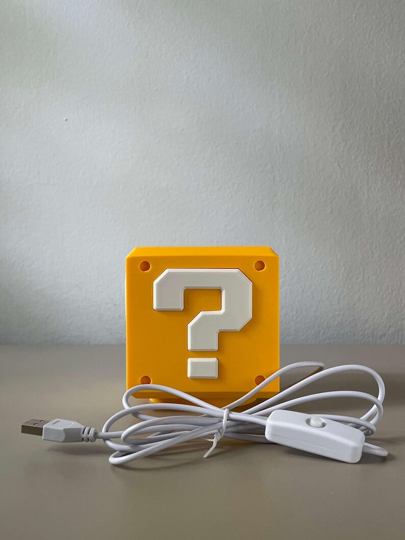 May include: A yellow cube-shaped lamp featuring a white question mark symbol on the front face. The lamp includes a white power cord with a USB connector and an inline toggle switch for operation, designed with a smooth, matte finish surface.