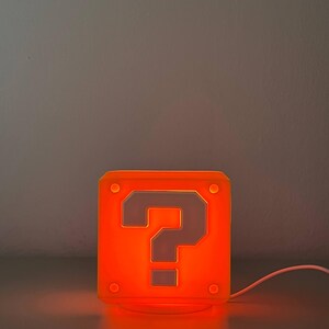 May include: A square, orange-glowing lamp shaped like a block with a prominent question mark symbol on the front face. The lamp features small circular indentations at each corner and is connected to a white power cord extending to the right side.