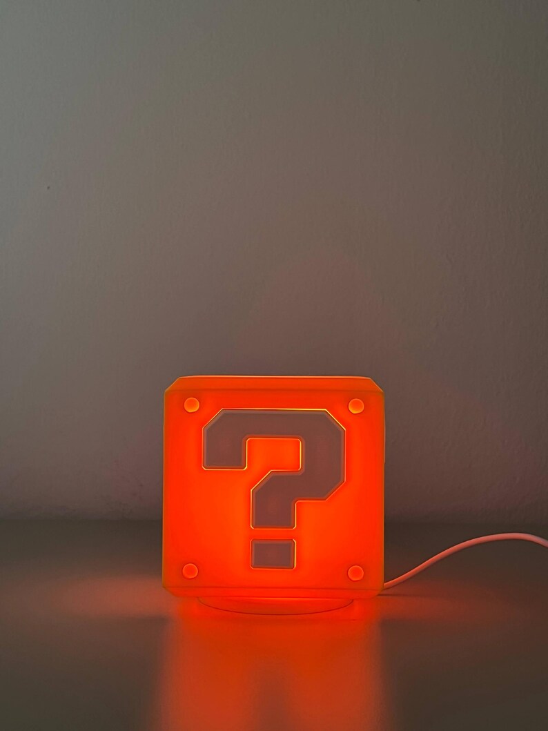 May include: A square, orange-glowing lamp shaped like a block with a prominent question mark symbol on the front face. The lamp features small circular indentations at each corner and is connected to a white power cord extending to the right side.