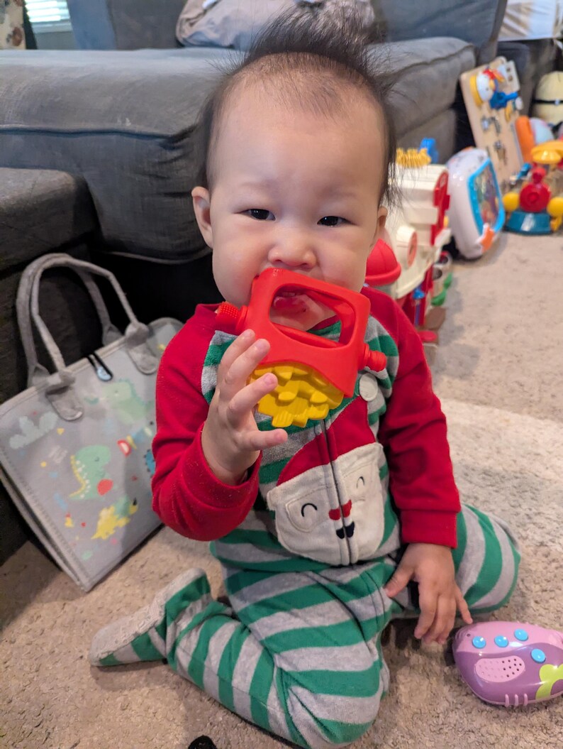 Pu&ograve; includere: Un bambino che indossa una tutina a righe rosse e verdi con un disegno di Babbo Natale sul davanti. Il bambino tiene in bocca un giocattolo rosso e giallo. Altri giocattoli sono visibili sullo sfondo.