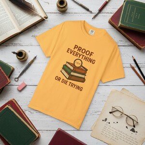 May include: Mustard yellow t-shirt with the text "PROOF EVERYTHING OR DIE TRYING" above a graphic of three books and a magnifying glass. The shirt is surrounded by books, pens, and other stationery.