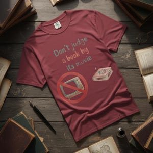 May include: A maroon t-shirt with the text "Don't judge a book by its movie" in teal and pink. The shirt features a graphic of a book and a crossed-out television. The shirt is surrounded by books and writing supplies.