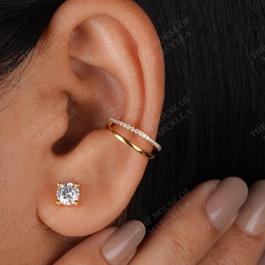 May include: A close-up of an ear adorned with gold jewelry. A gold ear cuff with a row of small, clear stones sits on the upper ear. A gold stud earring with a single, clear, round stone is in the earlobe.