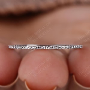 May include: A delicate, thin ring with a band of small, sparkling diamonds. The ring is set against a soft, blurred background. The House of Marcella is printed on the background.