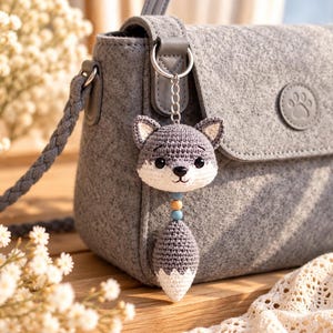 May include: A grey felt crossbody bag with a circular paw print emblem on the flap. Attached to the silver metal ring is a crocheted fox keychain featuring a grey and white head, a small blue and yellow bead, and a grey and white tail.