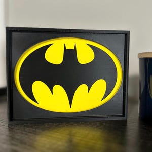 May include: A black framed Batman logo sign. The logo features a black bat silhouette on a yellow oval background. The sign is a 3D printed design, ideal for fans of the DC Comics superhero.