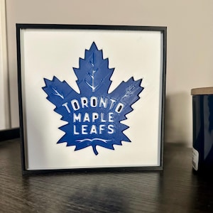 Puede incluir: Un marco cuadrado con un diseño de hoja de arce azul con el texto "TORONTO MAPLE LEAFS" en blanco. La obra de arte está sobre un fondo blanco dentro de un marco negro. La imagen es de temática deportiva.