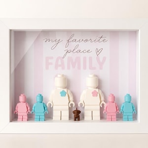 Può includere: Una scatola ombra con cornice bianca con sfondo a righe rosa e bianche. Il testo "my favorite place FAMILY" è visualizzato. Le figure Lego in rosa, blu e bianco sono disposte con una piccola statuina di cane marrone.
