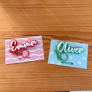 May include: Two personalized keychains, one red with the name "Emma" and the other green with "Oliver". Each keychain has a matching keyring and is displayed on a decorative card. The cards have the word "Lunique" printed on them.