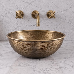May include: A round, hammered bronze vessel sink with a textured exterior finish. The sink is positioned on a white marble countertop with grey veining, accompanied by a wall-mounted bronze faucet and two matching cross-handle control valves.