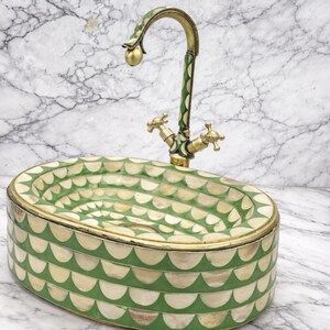 May include: An oval-shaped sink with a decorative green and white pattern. The sink has a brass faucet with green accents. The faucet has two cross-shaped handles. The sink is a unique bathroom fixture.