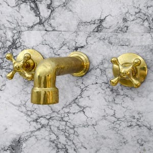 Può includere: Un set di rubinetti in ottone su sfondo marmoreo. Il set comprende un beccuccio curvo e due maniglie a forma di croce. Gli accessori hanno una finitura dorata lucida e il marmo presenta un motivo venato grigio e bianco.