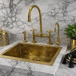 May include: A gold-coloured kitchen sink with matching tap and accessories. The sink is rectangular and hammered. A small plant in a gold pot and a decorative jar with a tassel are also present. The tap has a sprayer.