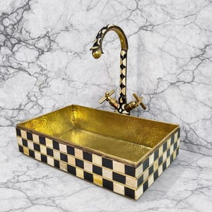 May include: A decorative bathroom sink with a gold interior and a black and white checkered exterior. The faucet is gold with black and white diamond patterns. The sink has a rectangular shape.