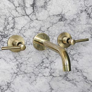 May include: A brass wall-mounted faucet set with a curved spout and two lever handles. The fixtures are set against a marble-patterned background, creating a contrast of textures and colors. The faucet has a vintage or antique aesthetic.
