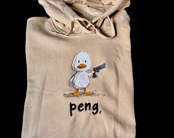 Könnte beinhalten: Beigefarbener Kapuzenpullover mit Kordelzug. Auf der Vorderseite ist eine gestickte Cartoon-Ente mit einer Pistole abgebildet, darunter das Wort "peng,". Der Hoodie ist aus weichem Material.