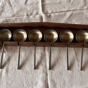 Op de afbeelding: Een set van zes metalen opscheplepels met ronde kommen, gepresenteerd op een donkerhouten rek. De lepels hebben lange, slanke handvatten en zijn netjes gerangschikt. Het rek is gemonteerd op een witte stoffen achtergrond.