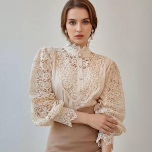 May include: A cream-coloured blouse with detailed lace sleeves and bodice. It has a high, lace-trimmed collar and pearl buttons. The long sleeves are puffy, with lace cuffs. The blouse is worn with a tan skirt. The blouse is a classic, elegant design.
