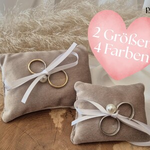 Könnte beinhalten: Zwei graue Ringkissen mit goldenen und silbernen Ringen, gebunden mit weißen Bändern und Perlen. Ein rosa Herz mit dem Text "2 Größen 4 Farben" im Hintergrund. Hochzeitsringhalter.