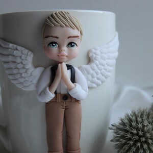 Puede incluir: Una taza de cer&aacute;mica blanca decorada con una figura de &aacute;ngel esculpida. El &aacute;ngel tiene cabello rubio, ojos azules y viste una camisa blanca, pantalones marrones y tirantes negros. Alas blancas est&aacute;n unidas a la taza.