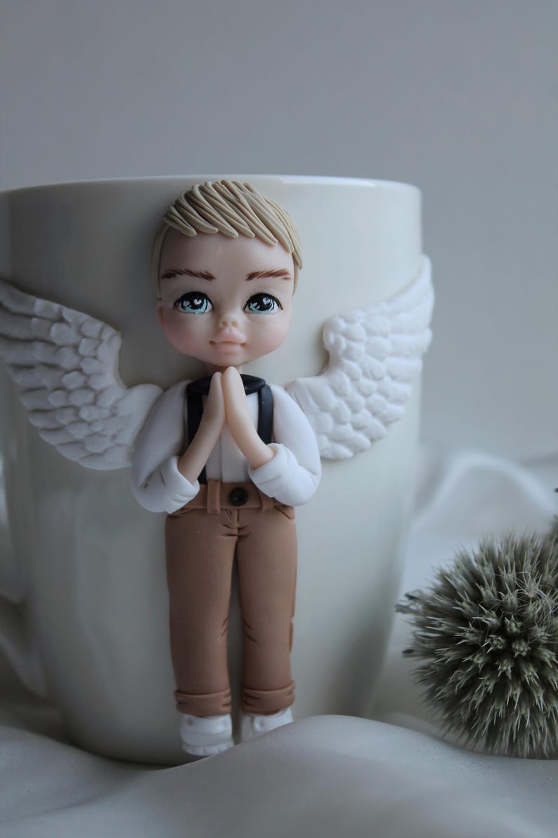 Puede incluir: Una taza de cer&aacute;mica blanca decorada con una figura de &aacute;ngel esculpida. El &aacute;ngel tiene cabello rubio, ojos azules y viste una camisa blanca, pantalones marrones y tirantes negros. Alas blancas est&aacute;n unidas a la taza.