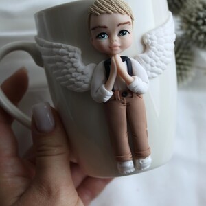 Puede incluir: Taza de cer&aacute;mica blanca con una figura de &aacute;ngel esculpida. El &aacute;ngel tiene cabello rubio, ojos azules y viste una camisa blanca, pantalones marrones y zapatos blancos. Alas blancas est&aacute;n adheridas a la taza.