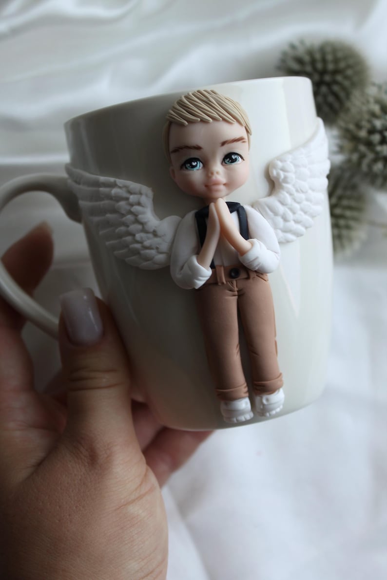 Puede incluir: Taza de cer&aacute;mica blanca con una figura de &aacute;ngel esculpida. El &aacute;ngel tiene cabello rubio, ojos azules y viste una camisa blanca, pantalones marrones y zapatos blancos. Alas blancas est&aacute;n adheridas a la taza.