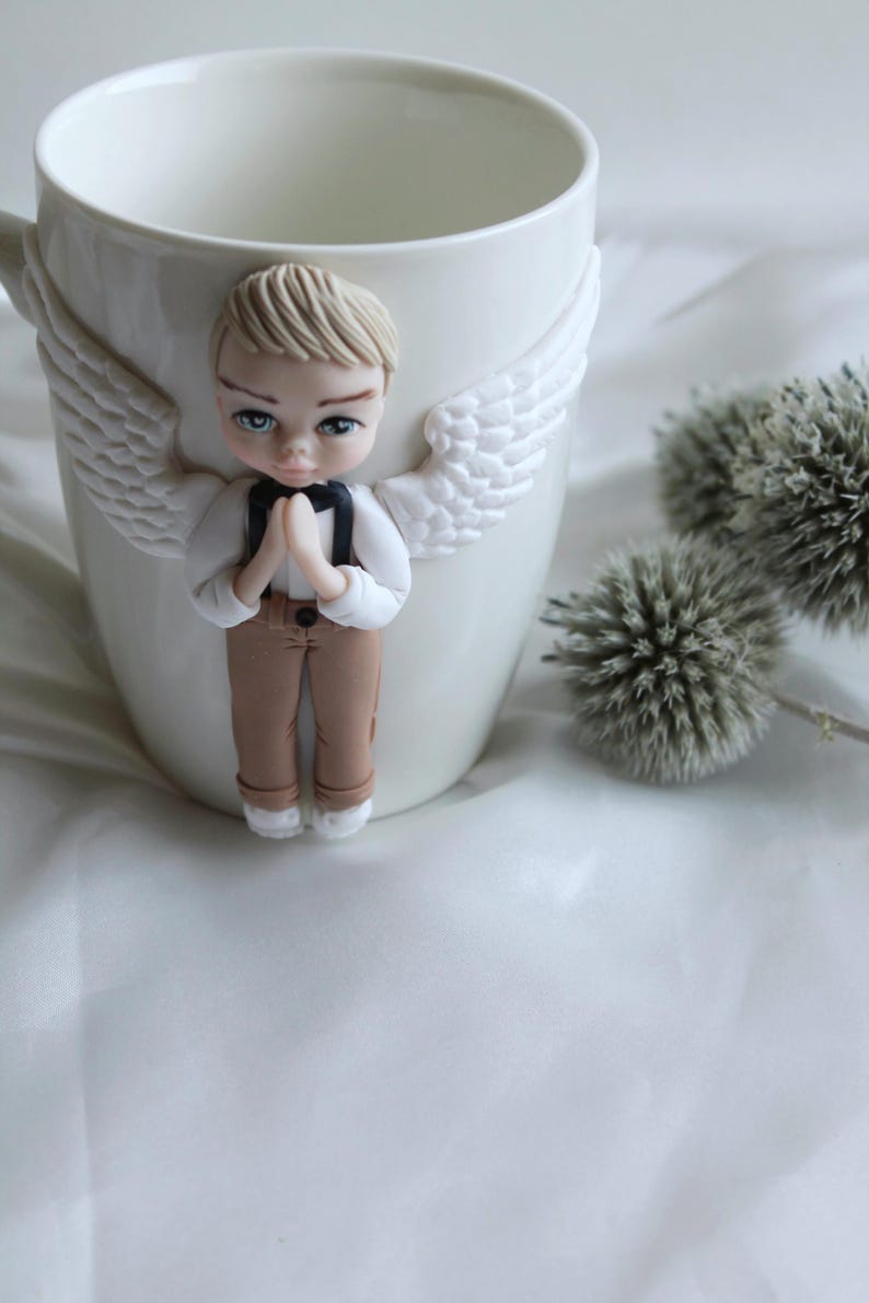 Puede incluir: Taza de cer&aacute;mica blanca con una figura de &aacute;ngel esculpida. El &aacute;ngel tiene cabello rubio, ojos azules y viste camisa blanca, pantal&oacute;n marr&oacute;n y pajarita negra. Alas blancas est&aacute;n adheridas a la taza. Cardos decorativos en el fondo.