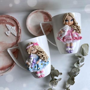 Puede incluir: Dos tazas de cerámica blanca decoradas con muñecas de arcilla polimérica. Una muñeca viste un vestido rosa y sostiene un conejito blanco. La otra muñeca viste un vestido azul y sostiene un unicornio. Bandejas decorativas rosas y blancas en el fondo.