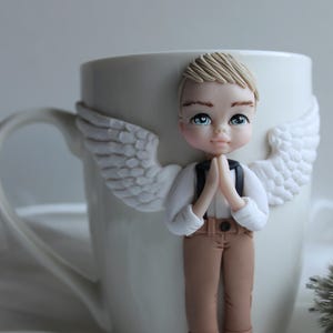 Puede incluir: Taza de cer&aacute;mica blanca decorada con una figura de &aacute;ngel ni&ntilde;o con cabello rubio, ojos azules y alas blancas. El &aacute;ngel lleva una camisa blanca, pantalones marrones y tirantes negros. La taza tiene un asa blanca.