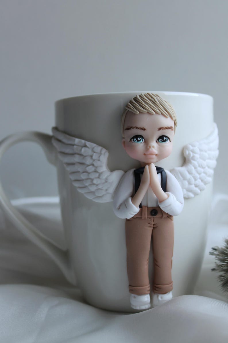 Puede incluir: Taza de cer&aacute;mica blanca decorada con una figura de &aacute;ngel ni&ntilde;o con cabello rubio, ojos azules y alas blancas. El &aacute;ngel lleva una camisa blanca, pantalones marrones y tirantes negros. La taza tiene un asa blanca.