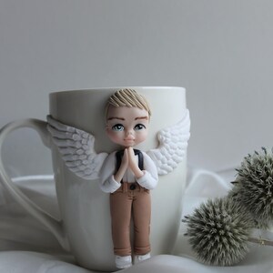 Puede incluir: Taza de cer&aacute;mica blanca con una figura decorativa de &aacute;ngel. El &aacute;ngel tiene cabello rubio, ojos azules y viste camisa blanca, pantal&oacute;n marr&oacute;n y alas blancas. El &aacute;ngel est&aacute; rezando con las manos juntas.