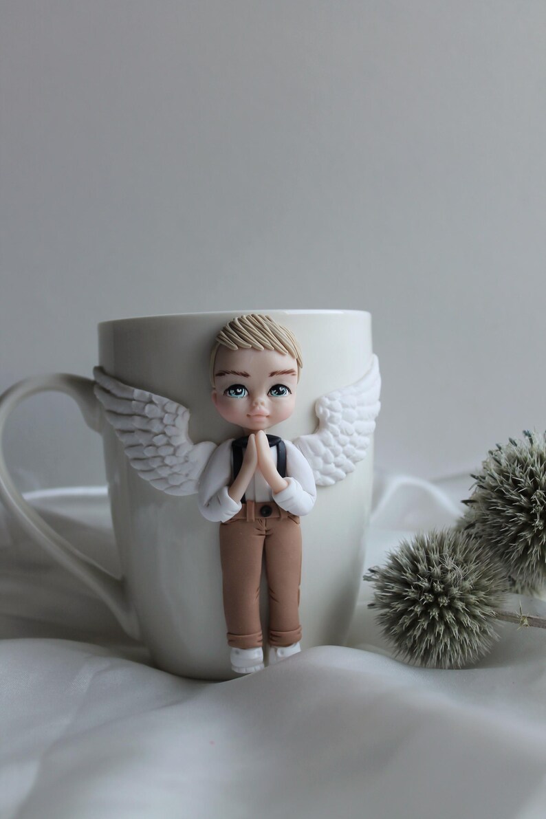 Puede incluir: Taza de cer&aacute;mica blanca con una figura decorativa de &aacute;ngel. El &aacute;ngel tiene cabello rubio, ojos azules y viste camisa blanca, pantal&oacute;n marr&oacute;n y alas blancas. El &aacute;ngel est&aacute; rezando con las manos juntas.