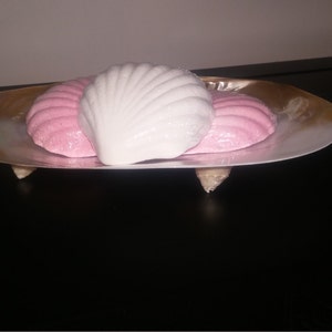 May include: Three decorative soap bars shaped like seashells, two are pink and one is white, displayed on a gold seashell dish.